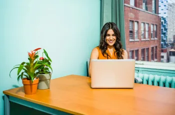 Confident woman in a bright office working on a laptop, surrounded by urban scenery and greenery.